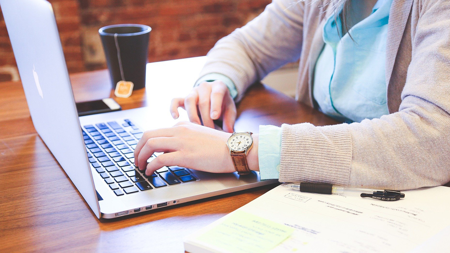 Instructor types on a laptop with a notebook pictured beside her on the table.