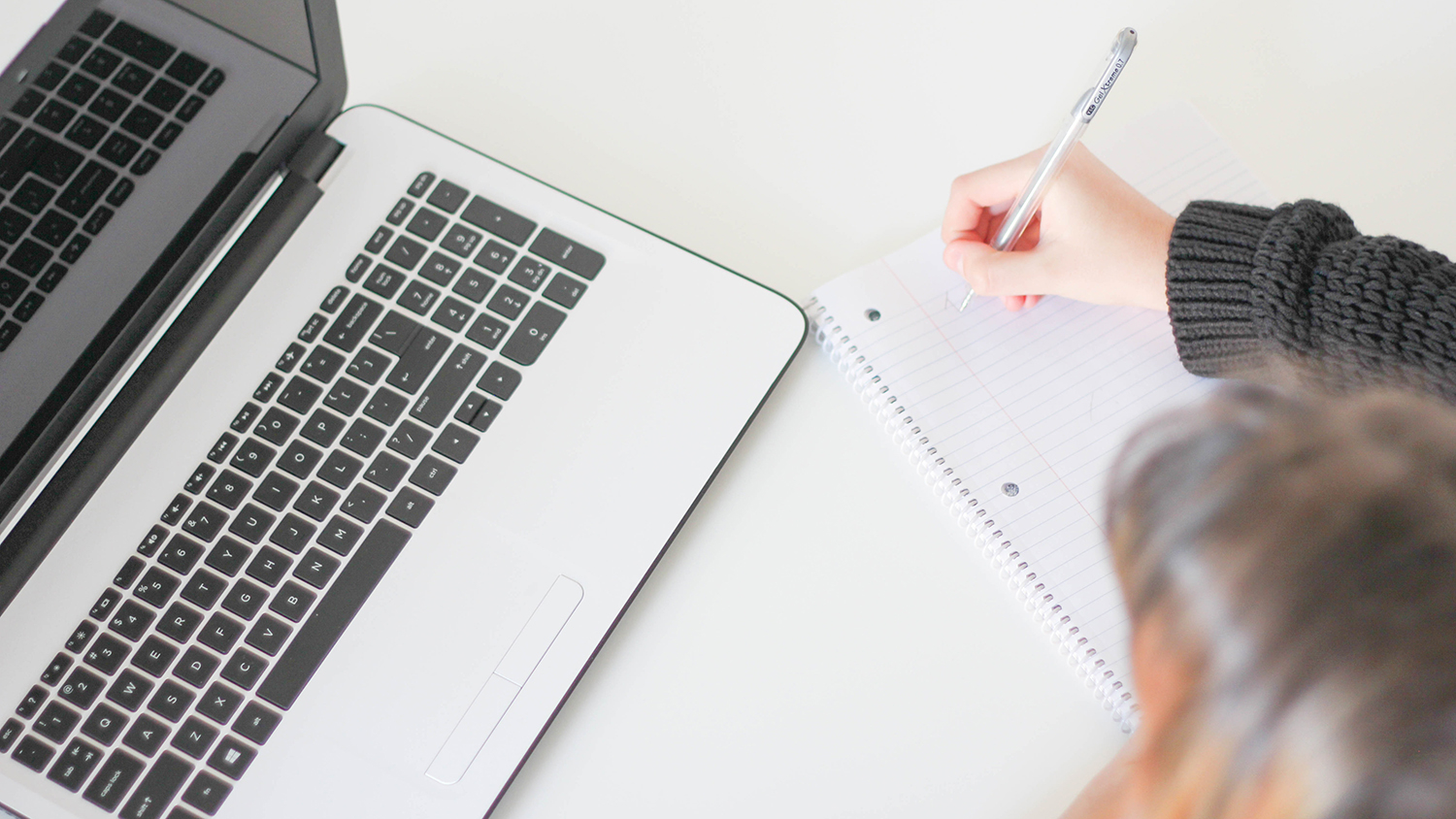 Photo of a laptop and a person writing on a piece of paper on a table. Photo is from Unsplash: jacqueline-kelly-PeUJyoylfe4