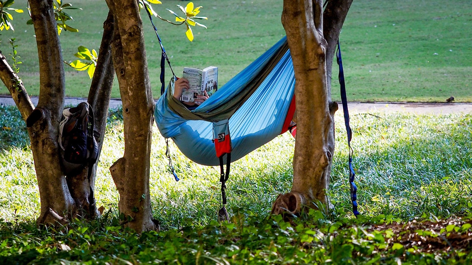 An NC State student reads in a hammock strung between two trees in a green space on campus.