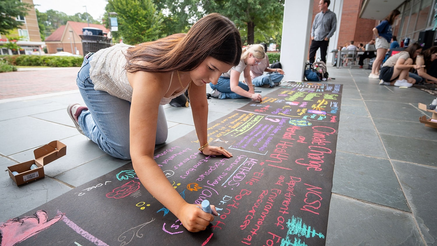Environmental Science student Ellie Boitnott works on a sidewalk chalk drawing of her research, titled "Climate Change and Mental Health!", during the Sidewalk Symposium.