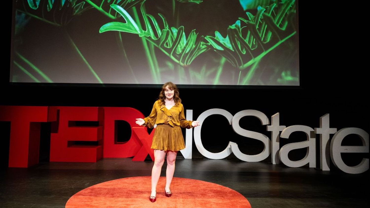 Student presents on a TEDxNCState stage, with a plant image projected behind her.